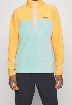 Personne portant un pull polaire avec des manches jaunes et le haut de la poitrine, un corps bleu clair, une patte de boutonnage rose et un pantalon noir.