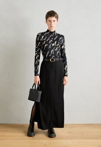 Femme aux cheveux courts portant un haut noir à manches longues avec motif, une jupe noire taille haute fendue, des bottes noires, une ceinture et tenant un petit sac à main noir.