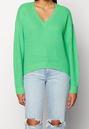 Persona que lleva un suéter de punto verde brillante con cuello en V y vaqueros rotos azul claro, de pie frente a un fondo liso.