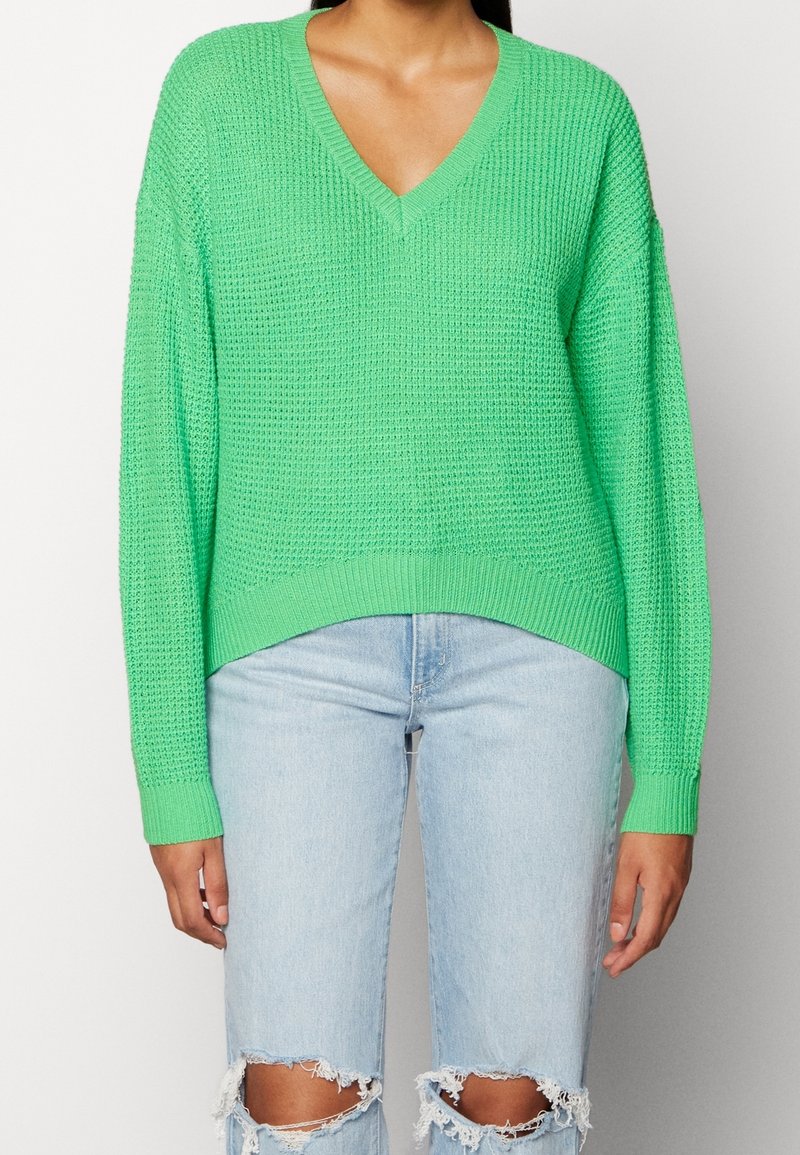 Personne portant un pull en maille vert vif à col en V et un jean bleu clair déchiré, debout devant un fond uni.