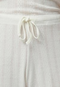 Pantaloni con vita elastica color crema con strisce verticali rosse. Presentano un laccio con coulisse, tessuto texturizzato con piccoli motivi a diamante.