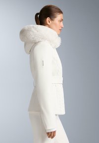 Giacca da sci bianca con cappuccio in pelliccia sintetica, dettaglio a maglia lavorata a cavo, vita con cintura e tasche laterali. Tessuto liscio e design moderno.