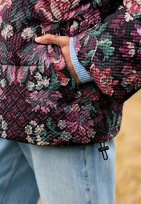 Schwarze gesteppte Steppjacke mit Blumenmuster in Grün, Pink und Lila. Mit gerippten blauen Bündchen und einer Tasche. Trägt man über Jeans.