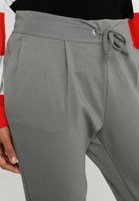 Lichtgrijze joggingsbroeken met een gladde textuur, elastische tailleband en een trekkoord. Heeft voorplooien en zijzakken.
