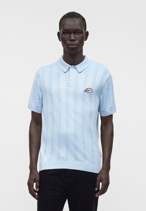 Homme portant un polo en maille bleu clair à manches courtes avec des rayures verticales texturées et un petit patch logo sur la poitrine, associé à un pantalon noir.
