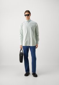 Camicia a bottoni verde chiaro, jeans blu, occhiali da sole neri e borsa a spalla nera. Presenta un piccolo logo sulla camicia e texture lisce.