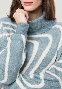 Frau trägt einen blau-weißen gemusterten Rollkragenpullover, mit der Hand, die in der Nähe ihres Kinns ruht, und sichtbaren Lippen.