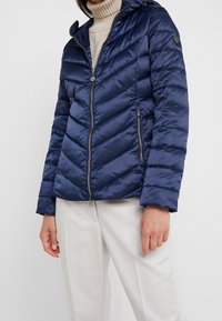 Mörkblå puffjacka med chevronmönster, dragkedjeslutning och huva. Slätt, glänsande tyg; har två fickor med dragkedja och en logotypdetalj.