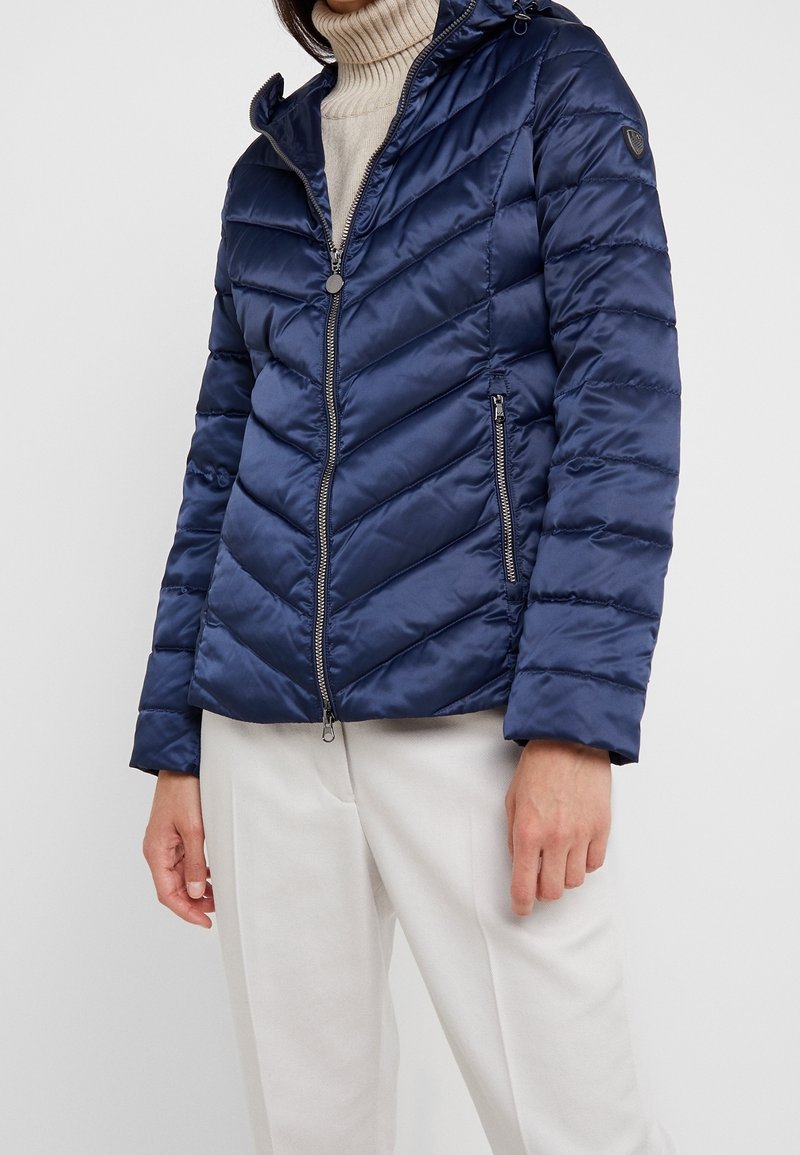 Mörkblå puffjacka med chevronmönster, dragkedjeslutning och huva. Slätt, glänsande tyg; har två fickor med dragkedja och en logotypdetalj.