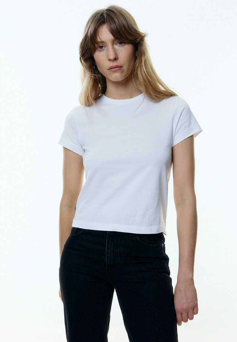 Weißes, kurzärmeliges T-Shirt mit rundem Ausschnitt und leicht gekürztem Saum. Kombiniert mit dunkelblauen Jeans. Glatte Stoffstruktur.
