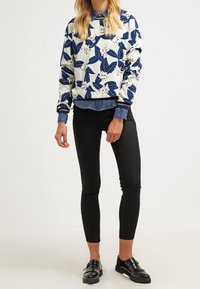 Felpa con motivo floreale blu su fondo crema, indossata sopra una camicia di denim. Abbinata a pantaloni neri attillati e scarpe nere lucide.