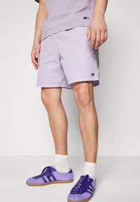 Světle fialové šortky vyrobené z lehkého materiálu, se bočním štítkem, spárované s purpurovými semišovými teniskami a bílými kotníkovými ponožkami.