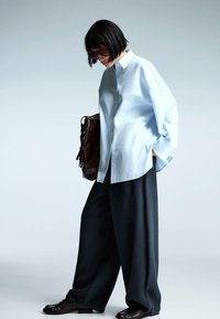 Jasnoniebieska, oversize koszula na guziki, ciemne spodnie z szerokimi nogawkami i brązowa torebka. Modelka nosi okulary przeciwsłoneczne, a tkanina ma gładkie wykończenie i luźny krój.