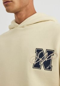 Beżowy hoodie z kapturem, wykonany z teksturowanej tkaniny. Zawiera haftowaną granatową literę "N" oraz wirujący akcent w kremowych nitkach. Prosty, casualowy fason.