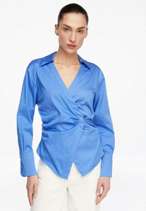 Kobieta z zaczesanymi do tyłu włosami, ubrana w długorękawową, jasnoniebieską bluzkę wiązaną i białe spodnie, stojąca na tle białego tła.