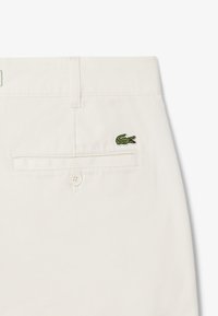 Pantaloni in cotone off-white con logo di coccodrillo ricamato in verde. Presentano una tasca posteriore con bottone e cuciture pulite.