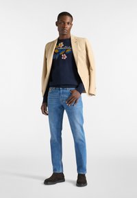 Beige kavaj över en marinblå tröja med blommig broderi, kombinerad med ljusblå jeans och bruna mockastövlar. Enkel, modern silhuett.