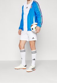 Blå atletisk jacka med flerfärgade ränder, vit tröja och shorts, svartvita fotbollssockor, vita fotbollsskor med orange accenter, fotboll.