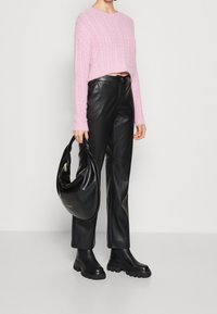 Suéter corto de punto texturizado en rosa claro, combinado con pantalones de piel sintética negros y botas de tobillo negras y voluminosas. Modelo sosteniendo un bolso negro.