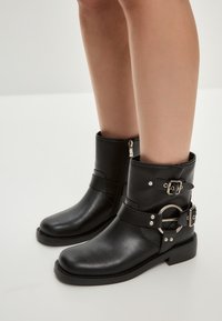 Botine negre, realizate din piele netedă, cu vârf rotunjit, fermoar pe lateral și curele decorative cu accente din metal.