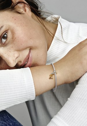 Silberarmband mit einem geknoteten Design, ausgestattet mit einem kleinen goldenen Bärenanhänger. Am Handgelenk getragen, mit einem strukturierten weißen Oberteil im Hintergrund.