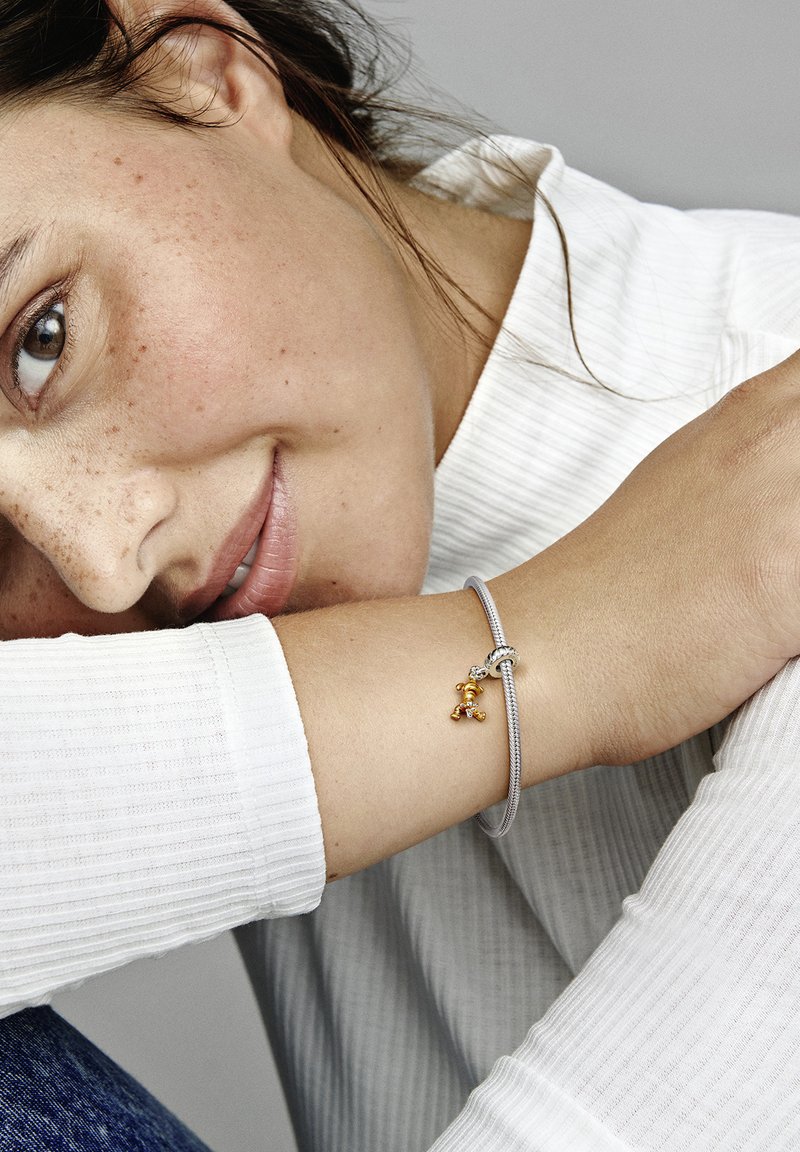 Silberarmband mit einem geknoteten Design, ausgestattet mit einem kleinen goldenen Bärenanhänger. Am Handgelenk getragen, mit einem strukturierten weißen Oberteil im Hintergrund.