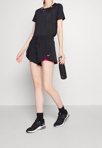 Top atlético negro de manga corta, combinado con pantalones cortos negros que tienen detalles en rosa. Modelo sosteniendo una botella de agua negra, vistiendo zapatillas negras.