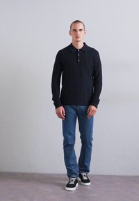 Bărbatul stă sprijinit de un perete simplu, purtând un pulover polo cu mânecă lungă, texturat, de culoare neagră, blugi și adidași negri cu tălpi albe.