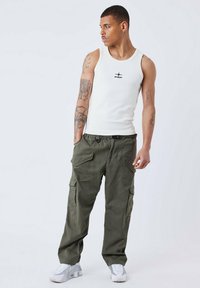 Top bianco senza maniche con logo nero, pantaloni cargo verde oliva con tasche laterali e scarpe da ginnastica bianche, su uno sfondo neutro.