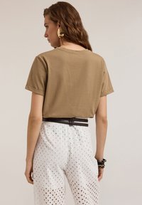 Camiseta de algodón marrón con mangas cortas, combinada con pantalones de pierna ancha blancos y perforados, y un cinturón marrón oscuro, visto desde la parte de atrás.