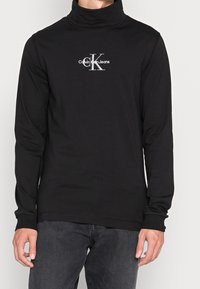 Mann trägt einen schwarzen Langarmrollkragenpullover mit weißem "Calvin Klein Jeans" Logo auf der Brust, kombiniert mit dunkelgrauen Jeans.