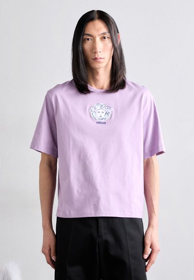 Fialové bavlněné tričko s volným střihem, které má na hrudi vyšívaný grafický design obličeje a text "VERSACE". Krátké rukávy a kulatý výstřih.