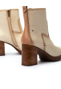 Beige leather ankle boots with a brown heel, featuring a side zipper, stitched detail, and metallic accents on the back.