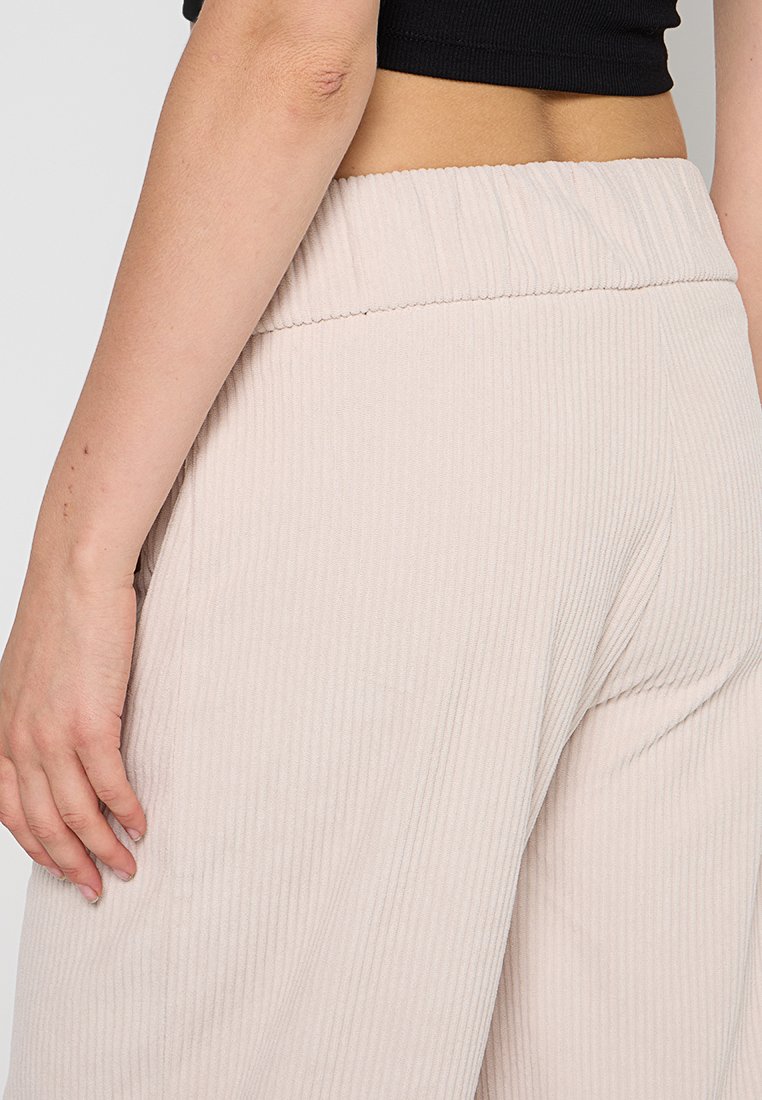 Pantaloni ampi a coste di un beige chiaro con vita elastica, texture liscia e vestibilità morbida, caratterizzati da un design casual e confortevole.