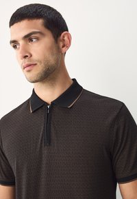 Zwart poloshirt met een subtiel golfpatroon, voorzien van een ritssluiting aan de voorkant en een contrasterende kraag. Zachte stof, korte mouwen, moderne pasvorm.