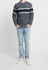 Persona vistiendo un suéter de punto a rayas en tonos azul marino y gris, jeans claros remangados y zapatillas blancas, de pie con las manos en los bolsillos.