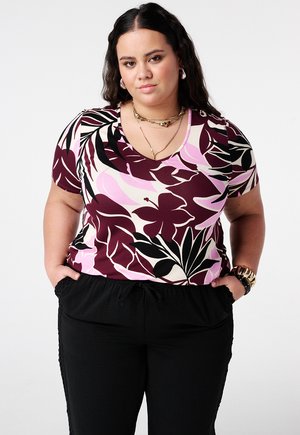 Vrouw met lang donker haar, gekleed in een korte mouw top met bloemenpatroon in wijnrood, roze en zwart, zwarte broek, gouden sieraden en een polshorloge, handen in de zakken.