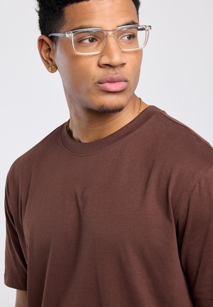 Young man wearing clear glasses, gold earrings, a gold chain, and a brown crewneck shirt looking to the side against a plain background.