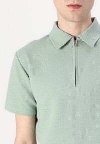 Svetlo zelena polo majica iz teksturiranega materiala, z ovratnikom in kratkimi rokavi, z sprednjim zadrgo.