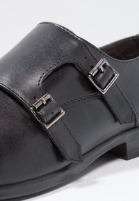 Chaussure en cuir noir à lanière avec deux boucles en argent et un petit talon sur fond blanc.