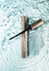 Máscara LH Cosmetics en un tubo plateado, parcialmente abierto, revelando un cepillo negro. La superficie tiene gotas y está situada sobre un fondo de agua.