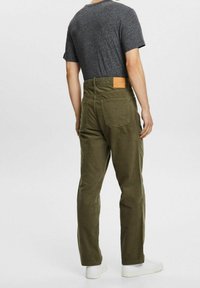 Pantalon en velours côtelé vert olive avec poches arrière, patch logo en cuir et jambes droites. Porté avec un t-shirt gris et des chaussures blanches.