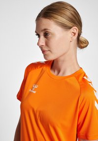 Ljusorange idrottströja med korta ärmar, med vita chevron-accenter på axlarna och en vit logotyp på bröstet. Slät textur.