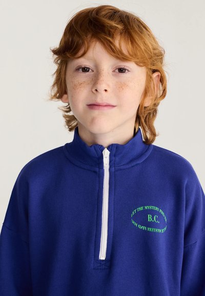 Enfant roux avec des taches de rousseur portant un pull zippé bleu avec un texte vert sur la poitrine, regardant droit devant avec une expression neutre.