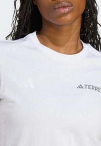 Camiseta de algodón blanca con cuello redondo, que presenta un logo gris "TERRE" y un diseño de tres franjas en el hombro. Textura suave, estilo sencillo.