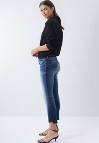 Vaqueros ajustados de color azul con un aclarado gradual hacia el dobladillo, con una cintura de altura media y longitud recortada. Combinados con una camisa negra de botones y sandalias con tiras.