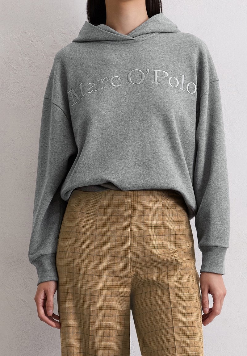 Sweat à capuche gris avec le texte « Marc O'Polo » associé à un pantalon marron à carreaux taille haute porté par une personne debout devant un mur neutre.