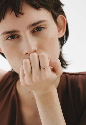Anillo de oro con tres piedras oscuras, llevado en un dedo. El sujeto tiene el cabello corto y oscuro y está posicionado contra un fondo neutro.