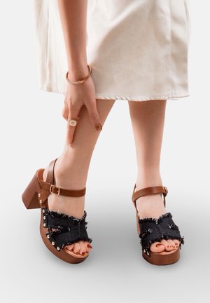 Pieds portant des sandales à talons brunes avec des lanières noires effilochées, une main avec une bague et un bracelet touchant la jambe au-dessus de la cheville, ourlet d'une robe blanche visible.