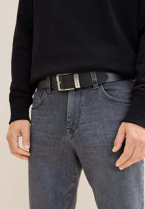 Ceinture en cuir noir avec une boucle en métal, portée sur un jean gris foncé. La ceinture a une texture lisse et une forme légèrement allongée.